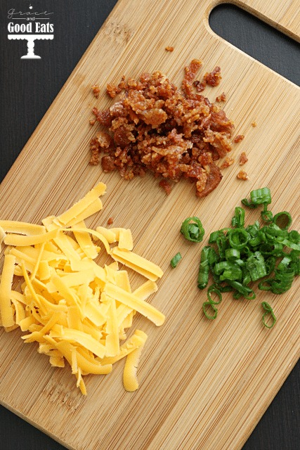 overhead view of crumbled bacon, shredded cheese, and chives on a wood cutting board 