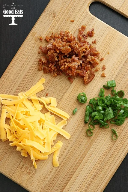 overhead view of crumbled bacon, shredded cheese, and chives on a wood cutting board 