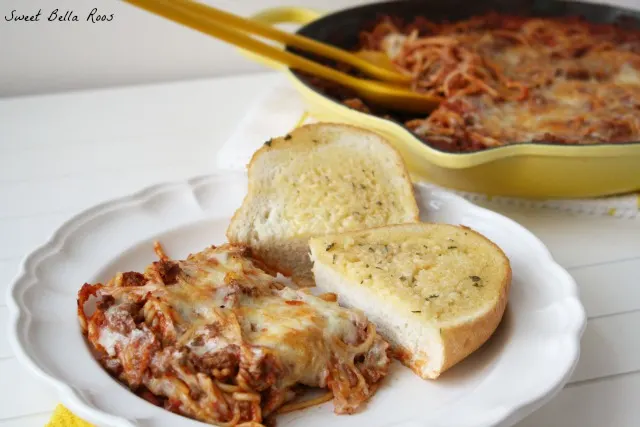 portion of cheesy baked spaghetti with garlic bread on a white plate. A skillet of spaghetti is in the background. 