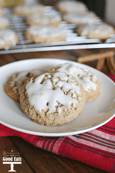 three iced oatmeal cookies on a white plate. A wire rack of cookies sits in the background. 