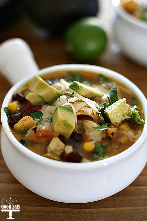 white soup crock bowl full of chicken tortilla soup with beans, corn, chicken, and tomatoes topped with cheese, corn chips, and avocados