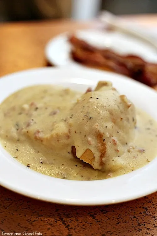 biscuits smothered in sausage gravy on a white plate with bacon in the background
