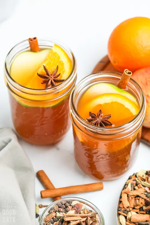 two glasses full of apple cider with oranges and spices beside them