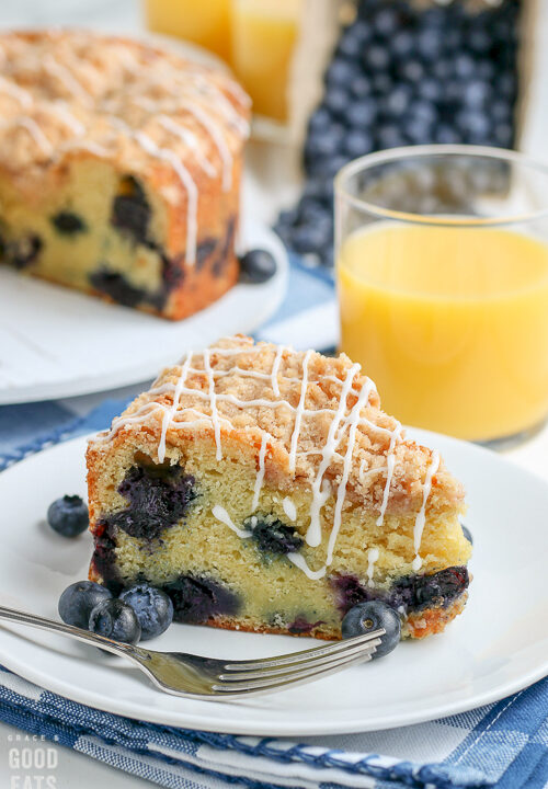 slice of blueberry coffee cake next to a fork and glass of orange juice