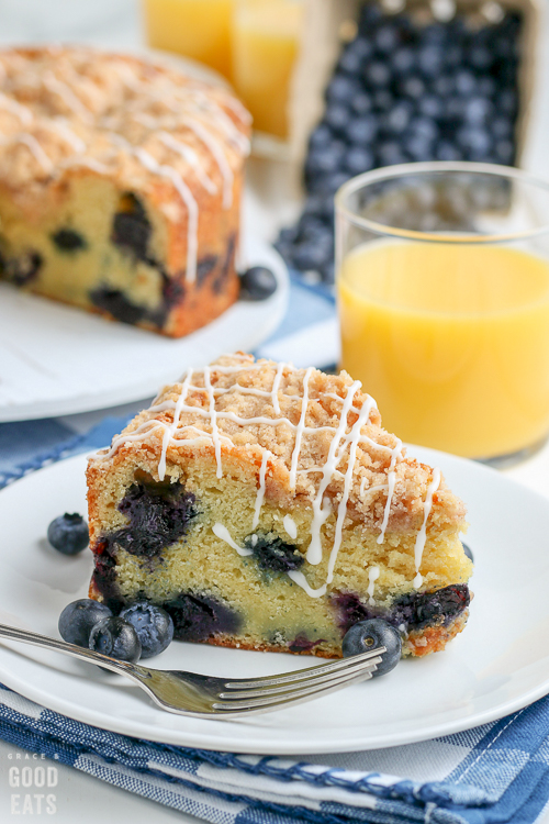 slice of blueberry coffee cake next to a fork and glass of orange juice