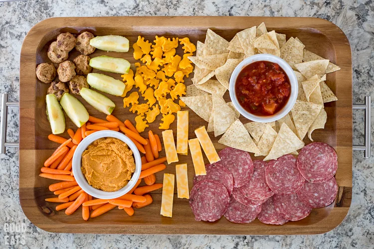 wooden tray with silver handles full of snacks like cheese sticks, salami, and crackers