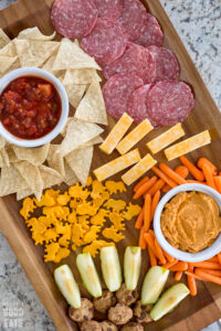 brown wooden tray with chips and salsa, veggies and hummus, apple slices, salami, cheese, and crackers