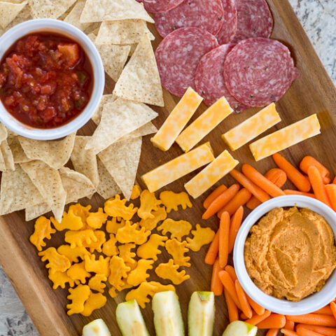 brown wooden tray with chips and salsa, veggies and hummus, apple slices, salami, cheese, and crackers