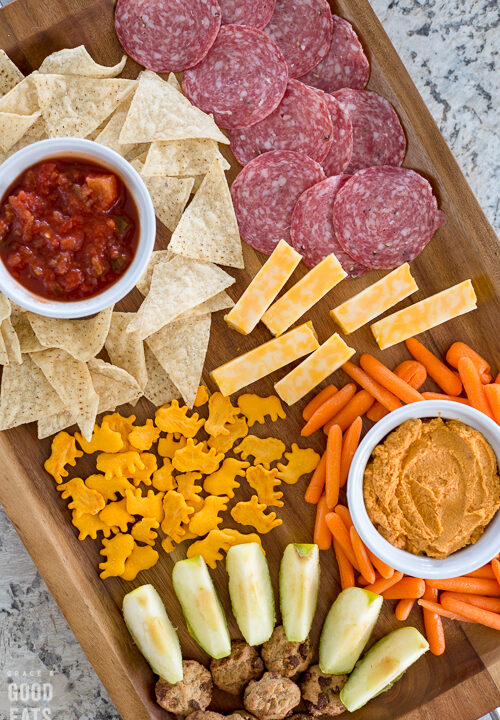 brown wooden tray with chips and salsa, veggies and hummus, apple slices, salami, cheese, and crackers