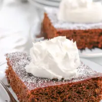 square slice of gingerbread cake on a white plate topped with whipped cream