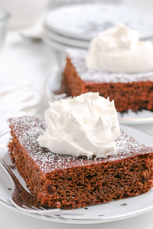 square slice of gingerbread cake on a white plate topped with whipped cream