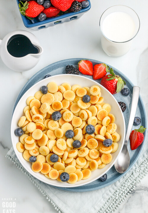 bowl of pancake cereal with blueberries next to a plate of strawberries and milk