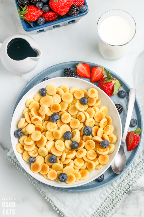bowl of pancake cereal with blueberries next to a plate of strawberries and milk